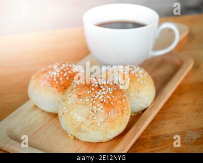 Frische hausgemachte Burger-Brötchen mit weißem Sesam auf einem kleinen Holztablett serviert mit heißem Kaffee auf Holztisch zum Frühstück. Drei leckere süße Sandwiches Stockfoto