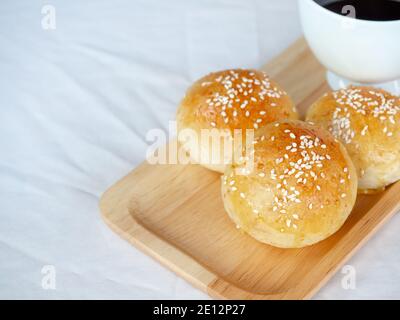 Frische hausgemachte Burger-Brötchen mit weißem Sesam auf Holztablett serviert mit heißem Kaffee auf weißem Tischtuch Hintergrund mit Copy Space. Drei leckere s Stockfoto