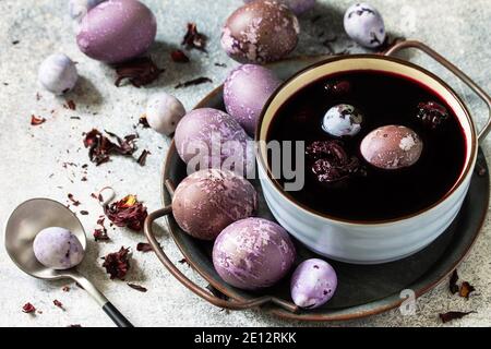 Natürliche Ostereier Farbstoff lila. Hausgemachte Eier werden mit natürlichem Eifarbstoff aus getrockneten Hibiskusblüten auf einer Tischplatte aus grauem Stein gemalt. Stockfoto