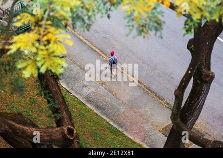 Luftaufnahme eines Mannes in roter Mütze, der einen Regenschirm hält und auf dem Bürgersteig mit dem Gefühl herbstlicher Blätter auf dem Boden läuft. Singapur Stockfoto