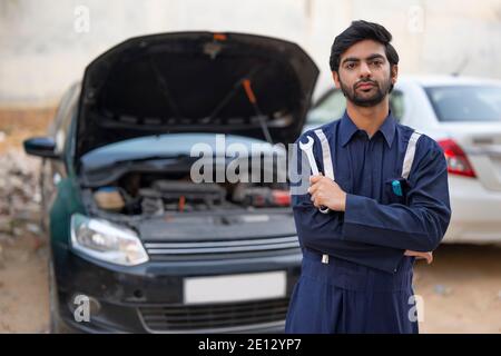 EIN JUNGER MECHANIKER, DER WERKZEUGE HÄLT, DIE VOR EINEM STEHEN ÖFFNEN SIE DAS FAHRZEUG Stockfoto