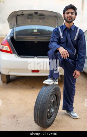 EIN JUNGER MECHANIKER STEHT SELBSTBEWUSST UND POSIERT MIT REIFEN Stockfoto