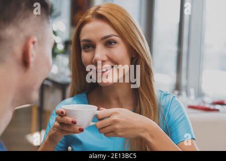 Nahaufnahme einer wunderschönen glücklichen rothaarigen Frau, die ihren Freund anlächelt, während sie zusammen Kaffee trinken. Schöne junge Frau genießen Morgenkaffee wi Stockfoto