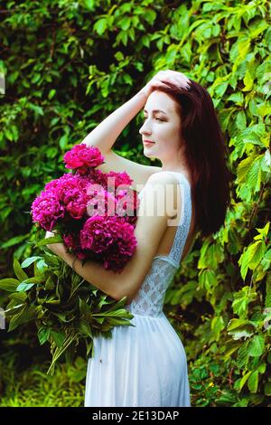 Porträt einer jungen attraktiven Frau im Frühlingsgarten mit blühenden rosa Pfingstrosen. Federhintergrund. Frauentag. Grußkarte. Stockfoto