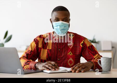 Afrikanischer Mann In Medizinischer Maske Und Traditionelle Kleidung Arbeiten An Heimbüro Stockfoto