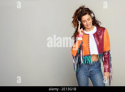 Trendige Frau in mehrfarbigen Jacke und Jeans stehen lächelnd mit Vergnügen, wie sie Musik auf Stereo-Kopfhörer gegen hört Eine weiße Wand mit CO Stockfoto