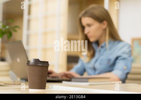 Selektive Konzentration auf eine Kaffeetasse auf dem Tisch Junge Frau arbeitet an ihrem Computer auf dem Hintergrund Copyspace Office Co-Working Workspace educati Stockfoto