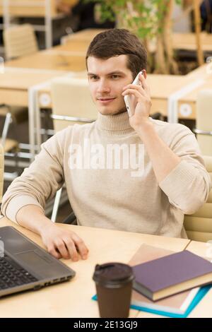 Vertikale Aufnahme eines hübschen jungen Mannes, der auf seinem spricht Das Telefon schaut weg und sitzt vor seinem Büro Die Computer-Technologie Konnektivität communi Stockfoto