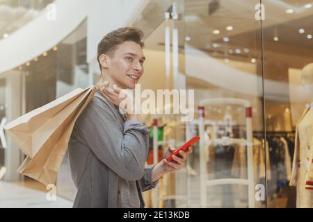 Junger, hübscher Mann, der sein Smartphone im Einkaufszentrum benutzt, Kopierraum. Fröhlicher männlicher Kunde genießt saisonale Verkauf in der Mall, trägt Ladenzeile Stockfoto