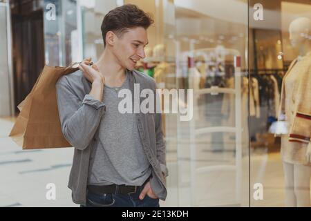 Attraktiver junger Mann, der lächelt, die Schaufenster des Ladens anschaut und im Einkaufszentrum spazieren geht. Schöner männlicher Kunde, der gerne im Einkaufszentrum einkauft, Kopie Stockfoto