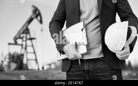 Beschnittene Nahaufnahme eines Ingenieurs, der seine Notiz und einen Stift in der einen Hand und einen weißen Helm in der anderen Hand hält, der im Ölfeld steht, Ölpumpenheber ist auf dem Hintergrund. Schwarzweiß-Bild Stockfoto