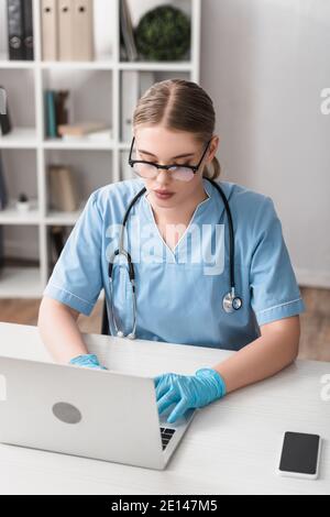 Junger Tierarzt in Brille und Latexhandschuhe tippen auf Laptop In der Nähe des Smartphones auf dem Schreibtisch Stockfoto