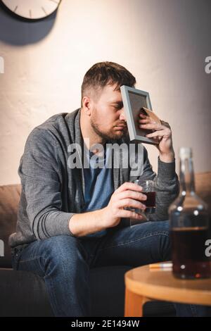 Deprimierter Mann, der das Gesicht mit einem Fotorahmen bedeckt, während er Glas hält Von Whiskey Stockfoto