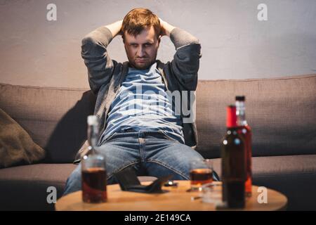 Betrunkener Mann, der neben dem Tisch sitzt, mit alkoholischen Getränken und leer Brieftasche im verschwommenen Vordergrund Stockfoto