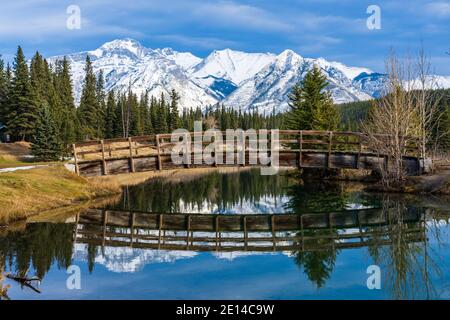 Holzbogen Fußgängerbrücke in Cascade Teiche Park im Herbst sonnigen Tag, schneebedeckten Mount Astley Reflexion auf der Wasseroberfläche. Banff National Park Stockfoto