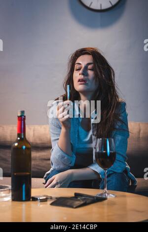 Betrunkene Frau hält Zigarette, während sie in der Nähe von Rotwein sitzt und Leere Brieftasche auf dem Tisch Stockfoto