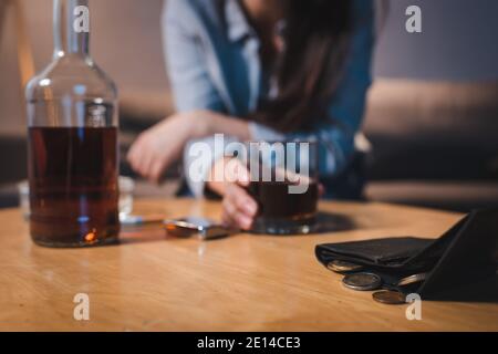 Beschnittene Ansicht der alkoholsüchtigen Frau, die ein Glas Whiskey in der Nähe der Brieftasche mit Münzen nimmt, verschwommener Hintergrund Stockfoto