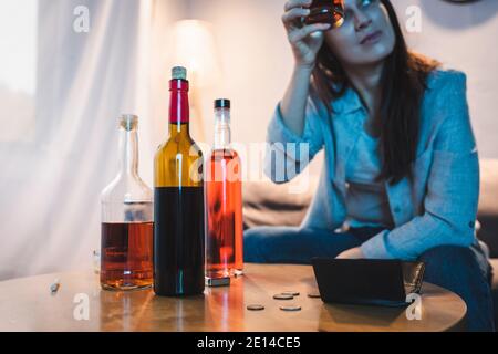 Betrunkene Frau hält Glas Alkohol in der Nähe von Flaschen, Münzen und leere Brieftasche auf dem Tisch, verschwommener Hintergrund Stockfoto