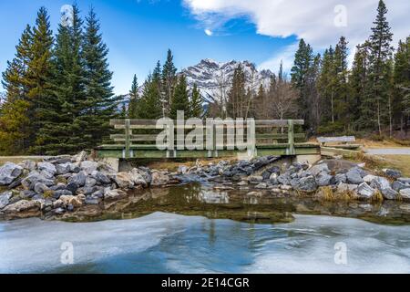 Holzsteg im Wald über gefrorene Eiswasseroberfläche im Winter. Schneebedeckter Berg im Hintergrund. Johnson Lake, Banff National Park, Can Stockfoto