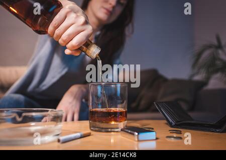 Beschnittene Ansicht einer Frau, die in der Nähe von Aschenbecher, Münzen und leerer Brieftasche Whiskey in Glas gießt, verschwommener Hintergrund Stockfoto