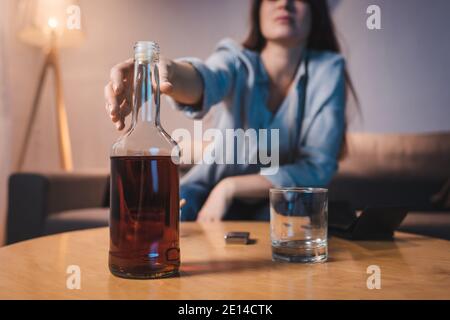 Beschnittene Ansicht einer alkoholischen Frau, die eine Flasche Whiskey vom Tisch nimmt, verschwommener Hintergrund Stockfoto