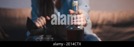 Beschnittene Ansicht der alkoholischen Frau mit Flasche und leerer Brieftasche auf verschwommenem Hintergrund, Banner Stockfoto