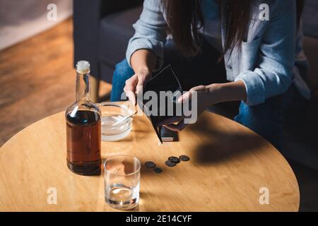 Beschnittene Ansicht der Frau, die leere Brieftasche in der Nähe von Flasche hält Whiskey und Münzen auf dem Tisch Stockfoto