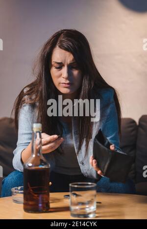 Betrunkene Frau hält leere Brieftasche in der Nähe von Glas und Flasche Whiskey bei verschwommenem Vordergrund Stockfoto