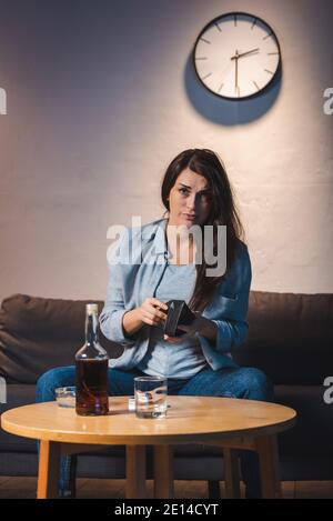 Betrunkene Frau, die die Kamera anschaut, während sie die leere Brieftasche in der Nähe hält Eine Flasche Whiskey auf dem Tisch Stockfoto