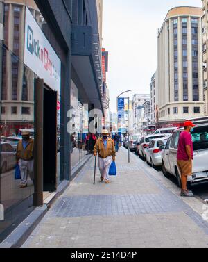 Kapstadt, Südafrika - 23/11/2020 Bewölkt Tag in Kapstadt. Menschen, die sich durch die Stadt bewegen. Älterer Mann mit Gesichtsmaske, der Lebensmittel trägt. Stockfoto