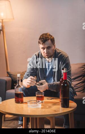 Betrunkener, frustrierter Mann, der die Kamera ansah, während er in der Nähe von Alkoholgetränken auf dem Tisch saß Stockfoto