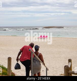 Camps Bay- Cape Town, South Africa - 23/11/2020 Junges Paar, das die Strandtreppen entlang geht, in Richtung leuchtend rosa Strandliegen. An einem warmen und bewölkten Tag. Stockfoto