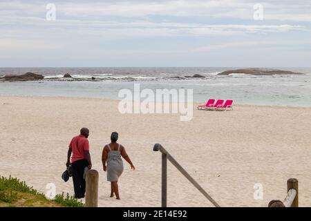 Camps Bay- Cape Town, South Africa - 23/11/2020 Junges Paar, das die Strandtreppen entlang geht, in Richtung leuchtend rosa Strandliegen. An einem warmen und bewölkten Tag. Stockfoto