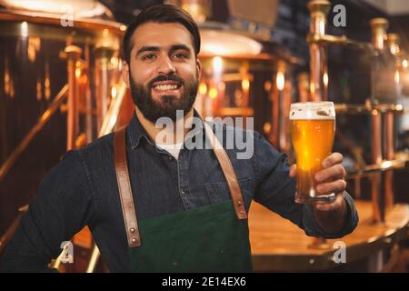 Fröhlicher bärtiger Bierbrauer lächelt freudig zur Kamera und hält ein Glas köstlichen, frisch gebrauten Bieres in der Hand. Fröhlicher Biermacher genießt die Arbeit an seinem b Stockfoto