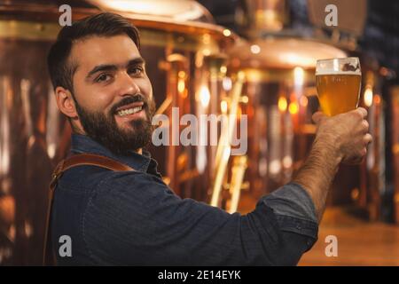 Glücklicher bärtiger Profi-Brauer lächelt der Kamera über die Schulter, hält ein Glas leckeres Bier, Kopierraum. Attraktive männliche Barkeeper-Position Stockfoto