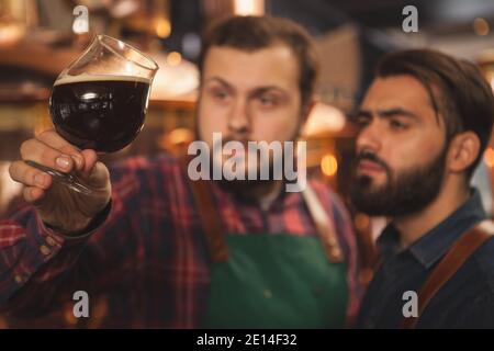 Selektive Konzentration auf ein Glas dunkles, köstliches Bier in den Händen professioneller Brauer. Zwei Bierhersteller untersuchen frisch gebrautes Bier und arbeiten bei ihnen Stockfoto