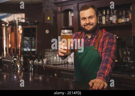 Fröhlicher Bartender mit Schürze, lächelnd zur Kamera, hält ein Glas Bier und posiert hinter der Theke. Freundlicher Brauer, der bei arbeitet Stockfoto