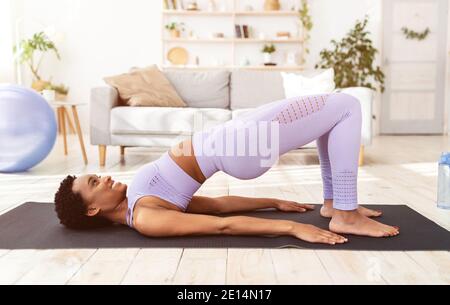 Sportliche schwarze Frau macht Übungen auf Yoga-Matte, stehend in Halbbrücke Pose drinnen, Seitenansicht Stockfoto