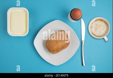 Direkt über Ansicht von Brötchen auf Teller, Butter, gekochtem Ei, Messer und Tasse Kaffee auf blauem Hintergrund, Frühstückskonzept Stockfoto