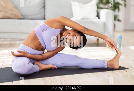 Happy Fit schwarze Dame streckt ihre Beine, macht Yoga-Posen oder Pilates auf Matte im Wohnzimmer Stockfoto