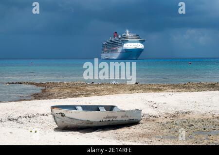 Der Blick auf ein altes kleines Boot auf der Grand Cayman Insel Seven Mile Beach und ein Kreuzfahrtschiff im Hintergrund unter dunklem regnerischen Himmel (Cayman Inseln). Stockfoto