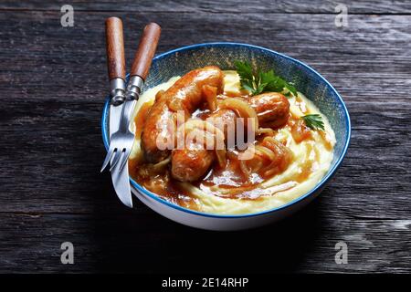 Hämmer und Maische traditionelle britische Gericht von Schweinefleisch Wurst mit Zwiebelsoße und Kartoffelpüree auf einem blauen Teller mit Besteck auf einem dunklen Holzhintergrund, Stockfoto