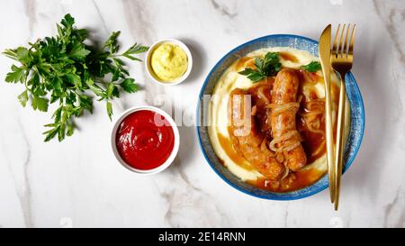 Traditionelle britische Gericht Knaller und Maische von Schweinefleisch Wurst mit Zwiebelsoße und Kartoffelpüree auf einem Teller mit goldenem Besteck, Ketchup und englischen Muste Stockfoto