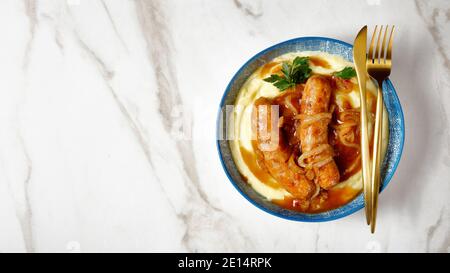 Hämmer und Maische traditionelle britische Gericht von Schweinefleisch Wurst mit Zwiebelsoße mit worcestershire-Sauce und Kartoffelpüree auf einem Teller mit goldenem Besteck Stockfoto