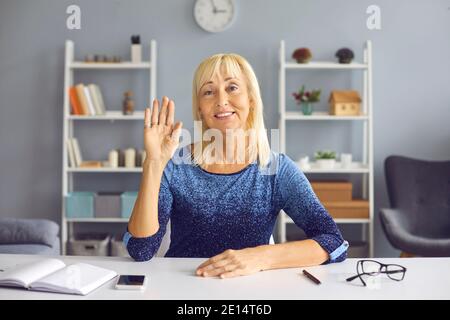 Erfolgreiche ältere Bloggerin winkt ihre Hand, während sie vor der Kamera an einem Tisch sitzt. Stockfoto
