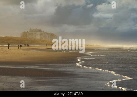 Goldenes Sonnenlicht scheint durch die brütenden dunklen Wolken, während die Menschen einen Spaziergang an einer winterlichen belgischen Küste machen. Die Wohnblöcke von Wenduine in West Stockfoto