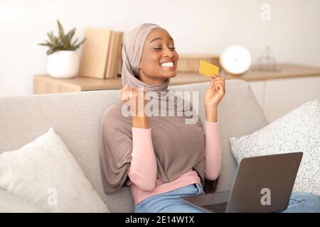 Fröhliche schwarze Dame in Hijab gestikulieren JA, halten Kreditkarte, Einkaufen im Online-Shop auf Laptop zu Hause Stockfoto