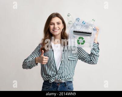 Lächelnde Frau hält Papierkorb mit Plastikflaschen und zeigt Daumen hoch Stockfoto