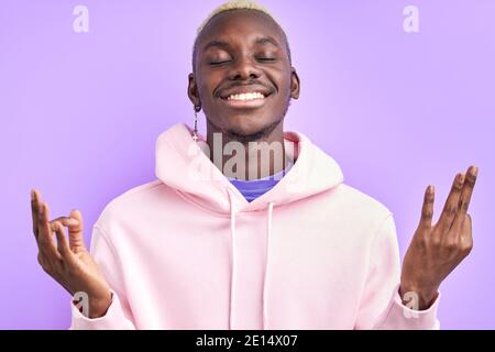 Optimistischer afrikanischer Mann hält ruhig, steht in Yoga-Pose. Schöner Mann in rosa Casual tragen Lächeln, mit geschlossenen Augen Stockfoto