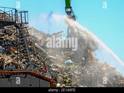 Bagger mit Krangreifer arbeitet auf einem mit Wasser gesprinklerten Schrottplatz im Hamburger Hafen Stockfoto